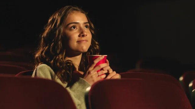 A young woman with long curly brown hair sits in a dark movie theater on a deep…